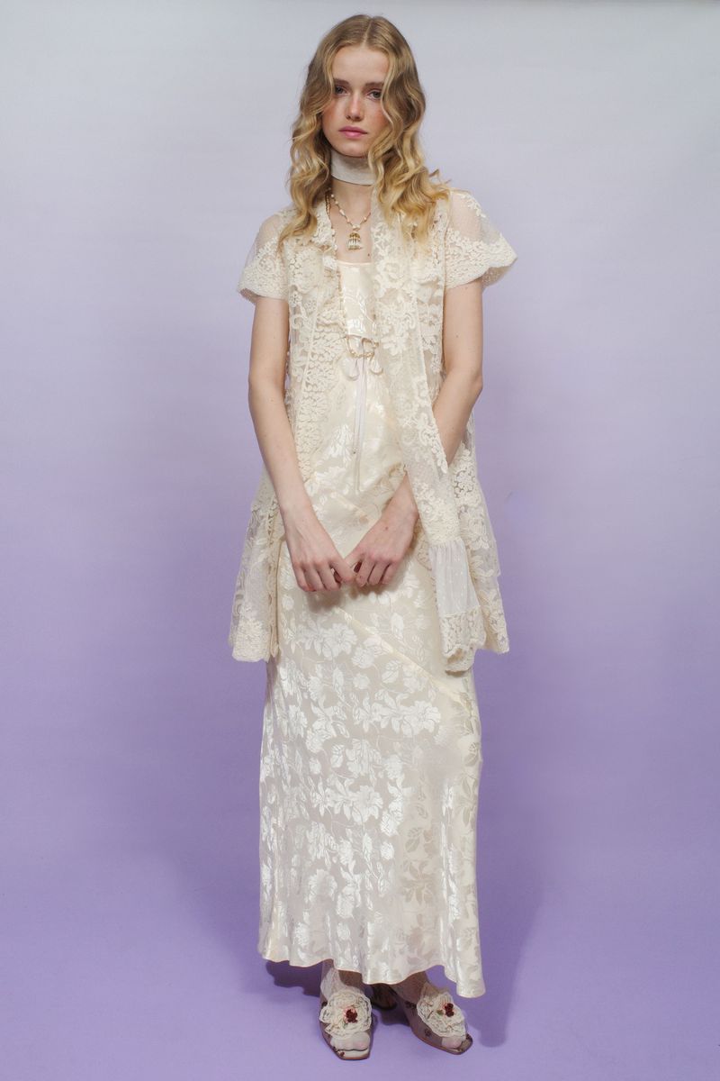Model standing in a layered cream floral lace dress and embroidered mules against a lavender seamless studio backdrop.