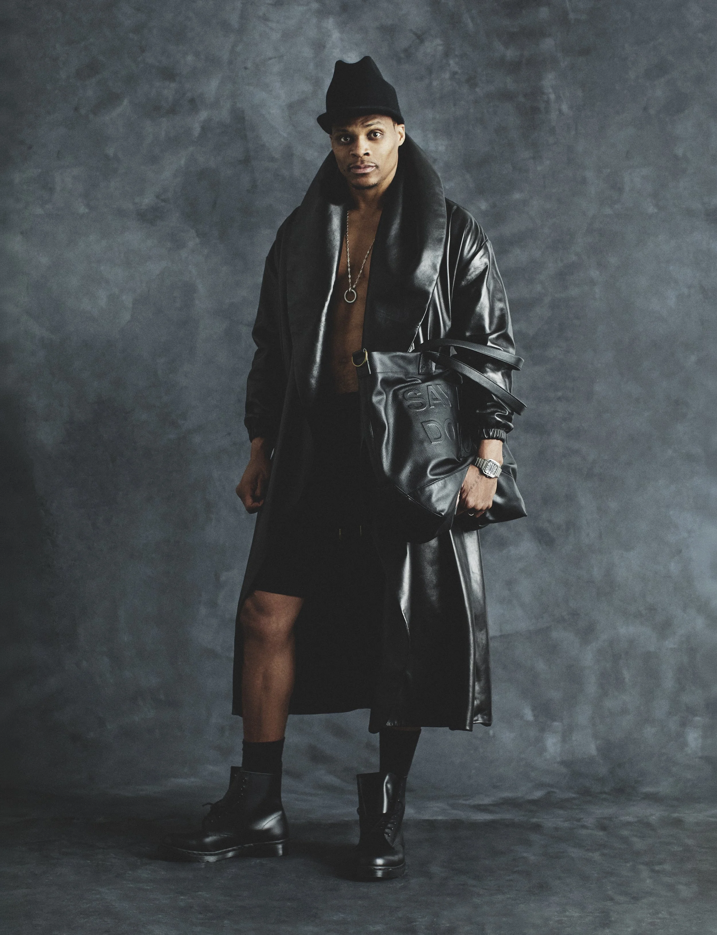 Russell Westbrook in a black leather trench coat, hat, and combat boots against a textured dark grey studio backdrop.