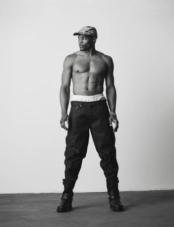 Russell Westbrook standing shirtless wearing dark denim pants, black boots, and a cap against a white studio background.