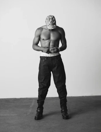 Shirtless Russell Westbrook looking down wearing dark jeans, combat boots, and a patterned cap against a neutral studio backdrop.