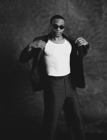 Russell Westbrook in sunglasses, a white tank top, and an open dark blazer against a mottled studio backdrop.