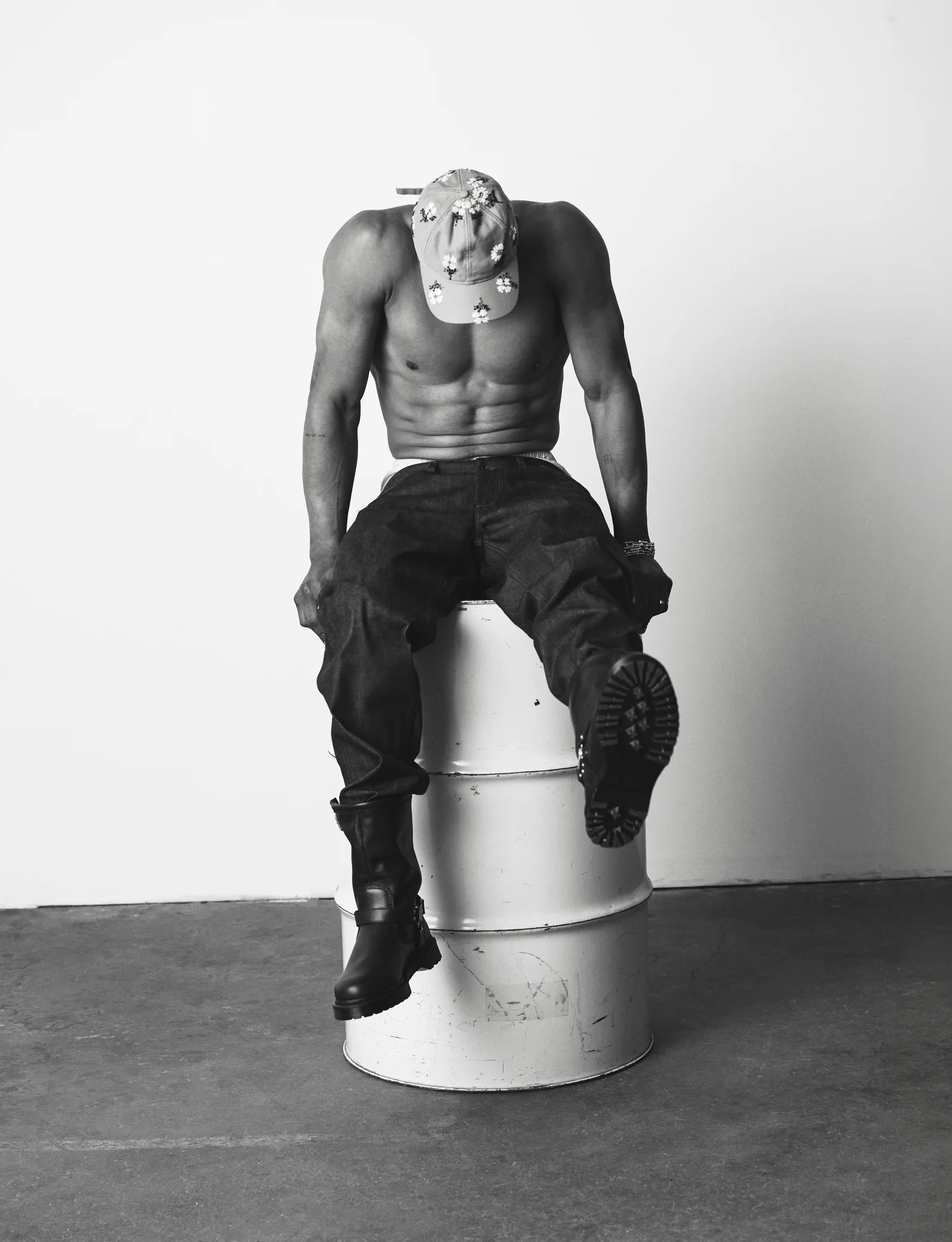 Shirtless man wearing dark cargo trousers, boots, and a patterned cap seated on a white oil drum against a studio backdrop.