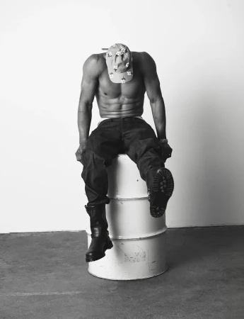 Shirtless man wearing dark cargo trousers, boots, and a patterned cap seated on a white oil drum against a studio backdrop.