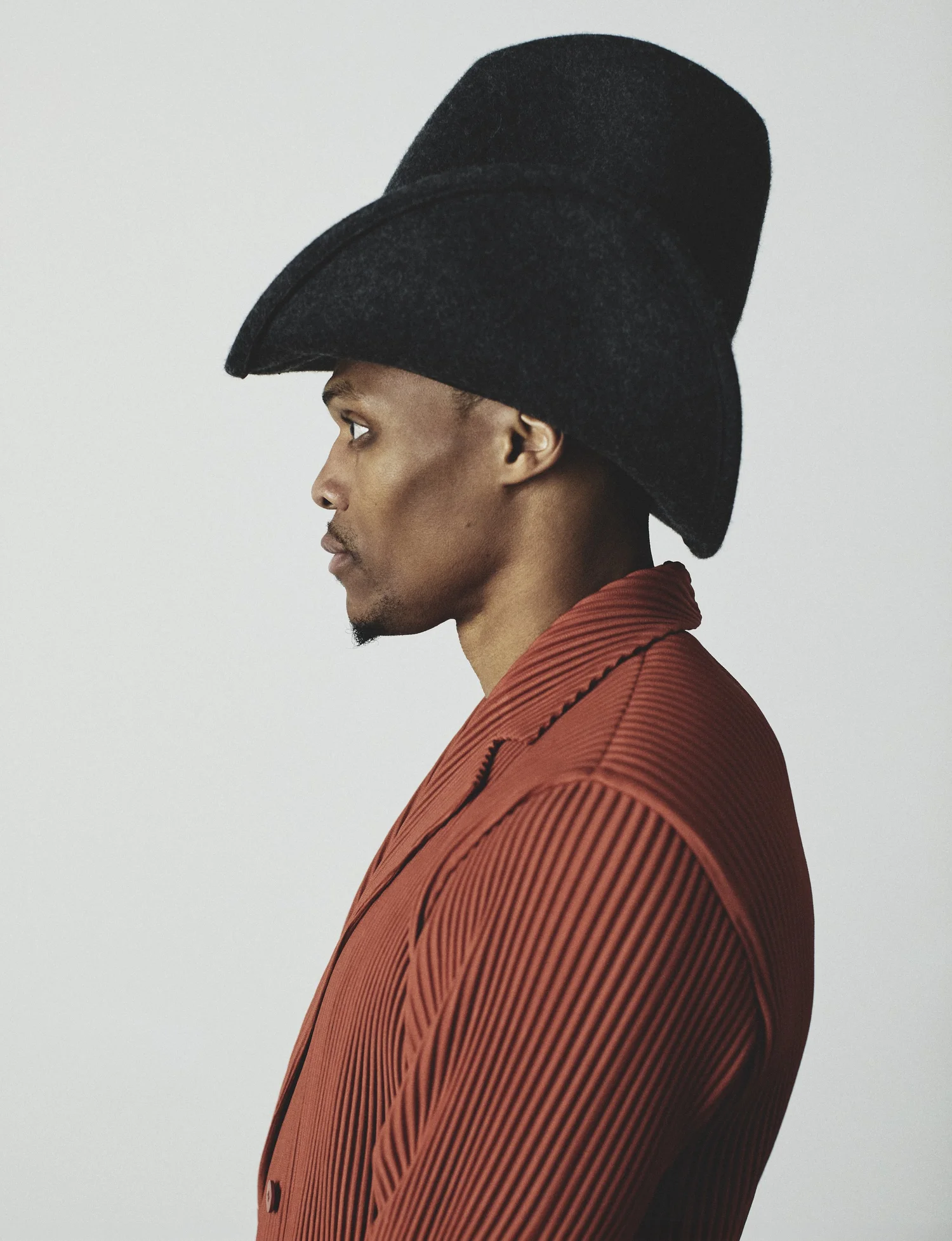 Profile portrait of Russell Westbrook wearing an exaggerated black felt hat and a pleated rust jacket against a light background.