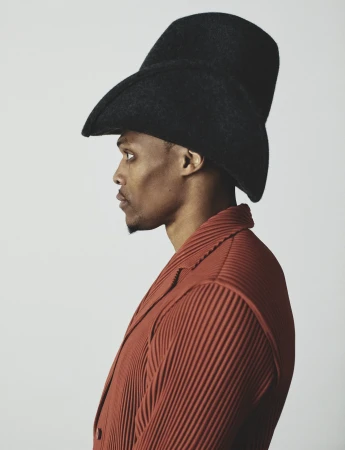 Profile portrait of Russell Westbrook wearing an exaggerated black felt hat and a pleated rust jacket against a light background.
