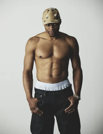 Russell Westbrook posing shirtless in a floral tan baseball cap and dark wide-leg trousers against a white seamless backdrop.