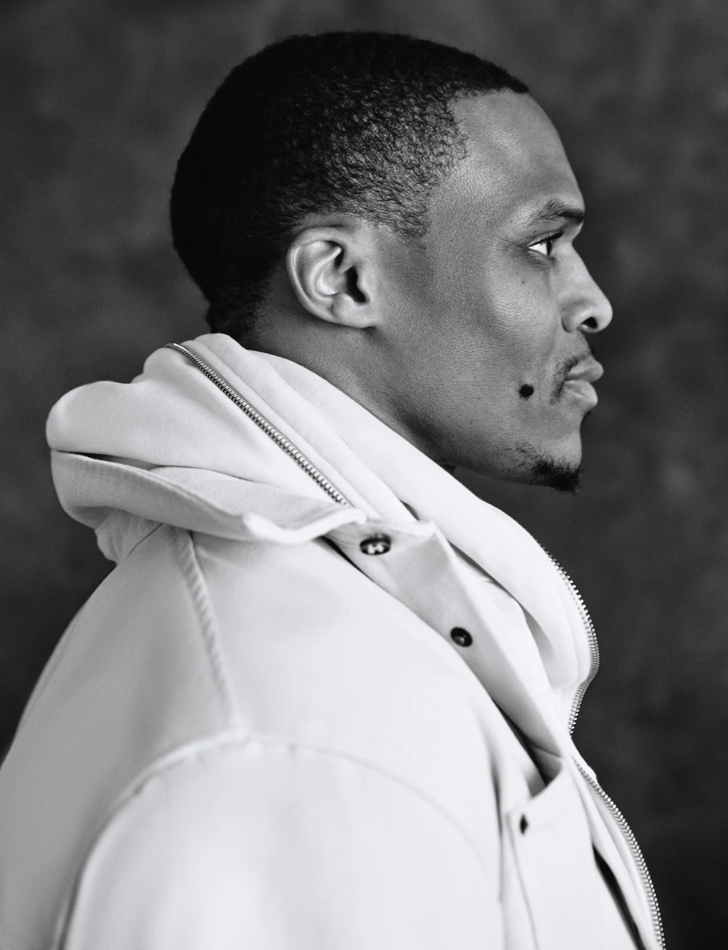 Black and white profile portrait of Russell Westbrook wearing a light hooded jacket against a dark textured backdrop.
