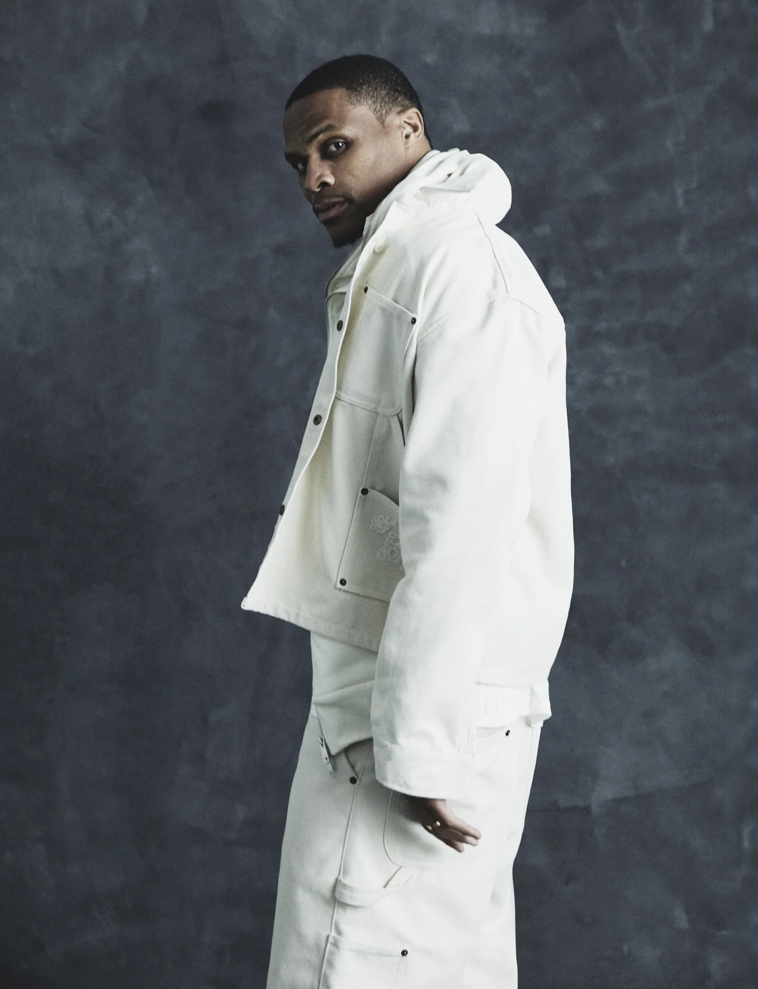 Russell Westbrook posing in an off-white denim workwear jacket and trousers against a dark textured backdrop.