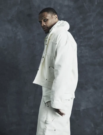 Russell Westbrook posing in an off-white denim workwear jacket and trousers against a dark textured backdrop.