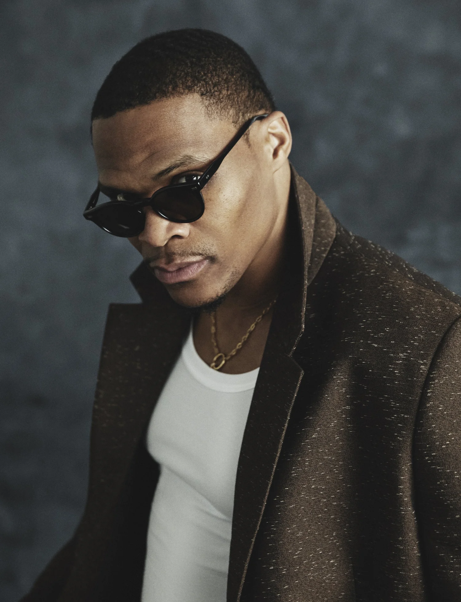 Russell Westbrook wearing dark sunglasses, a white tank top, and a textured brown coat in an editorial portrait.