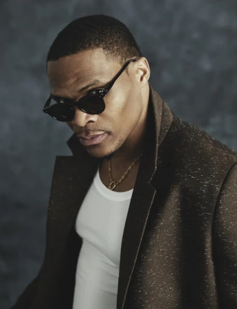 Russell Westbrook wearing dark sunglasses, a white tank top, and a textured brown coat in an editorial portrait.