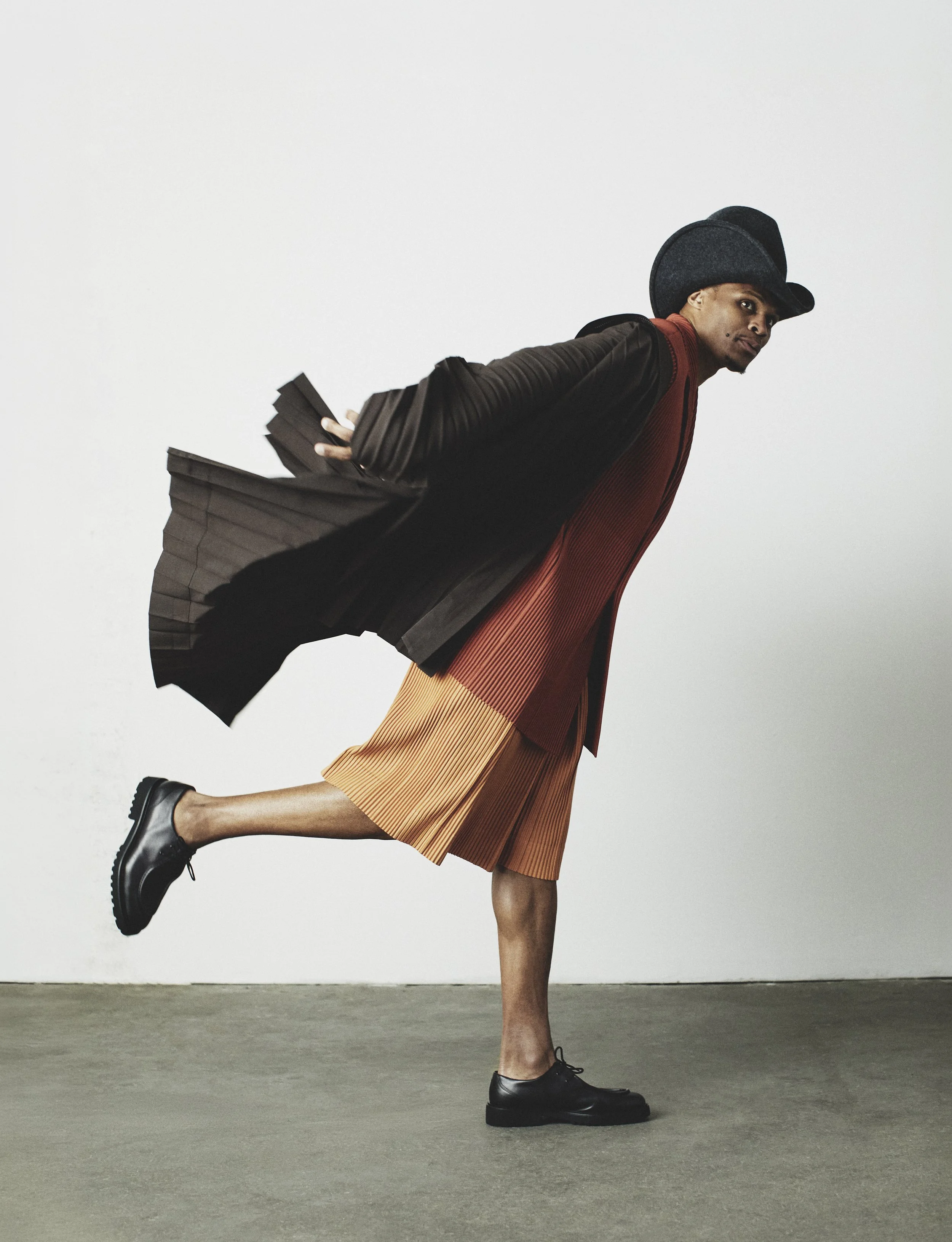Russell Westbrook wearing a pleated coat and hat in motion against a neutral studio background
