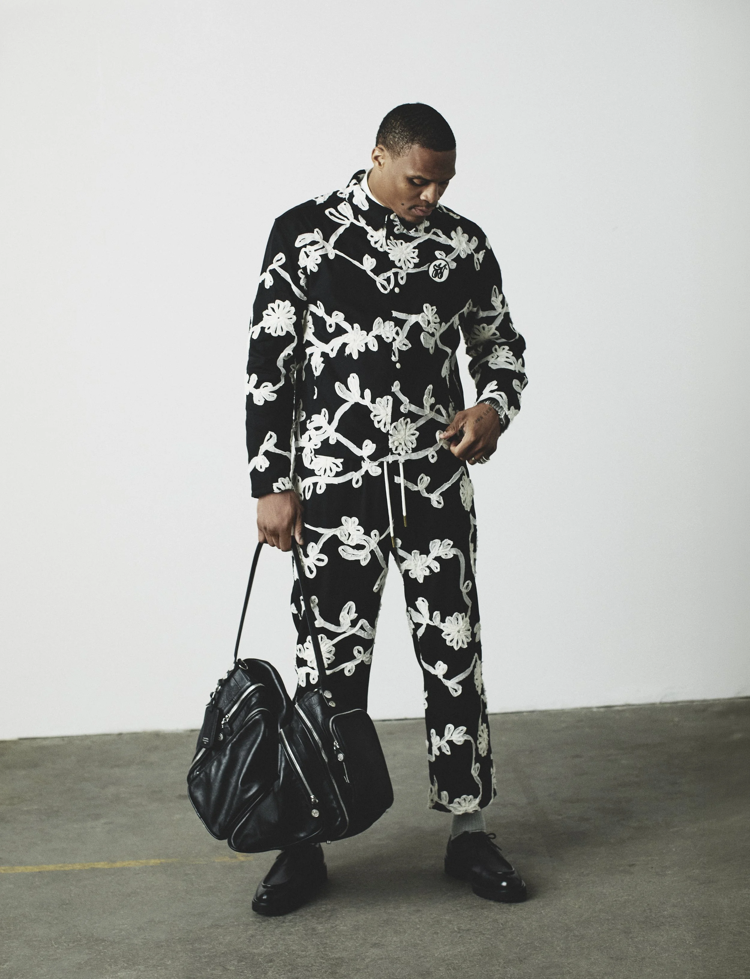 Russell Westbrook standing in a black and white embroidered two-piece set holding a black leather bag against a plain wall.