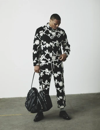 Russell Westbrook standing in a black and white embroidered two-piece set holding a black leather bag against a plain wall.