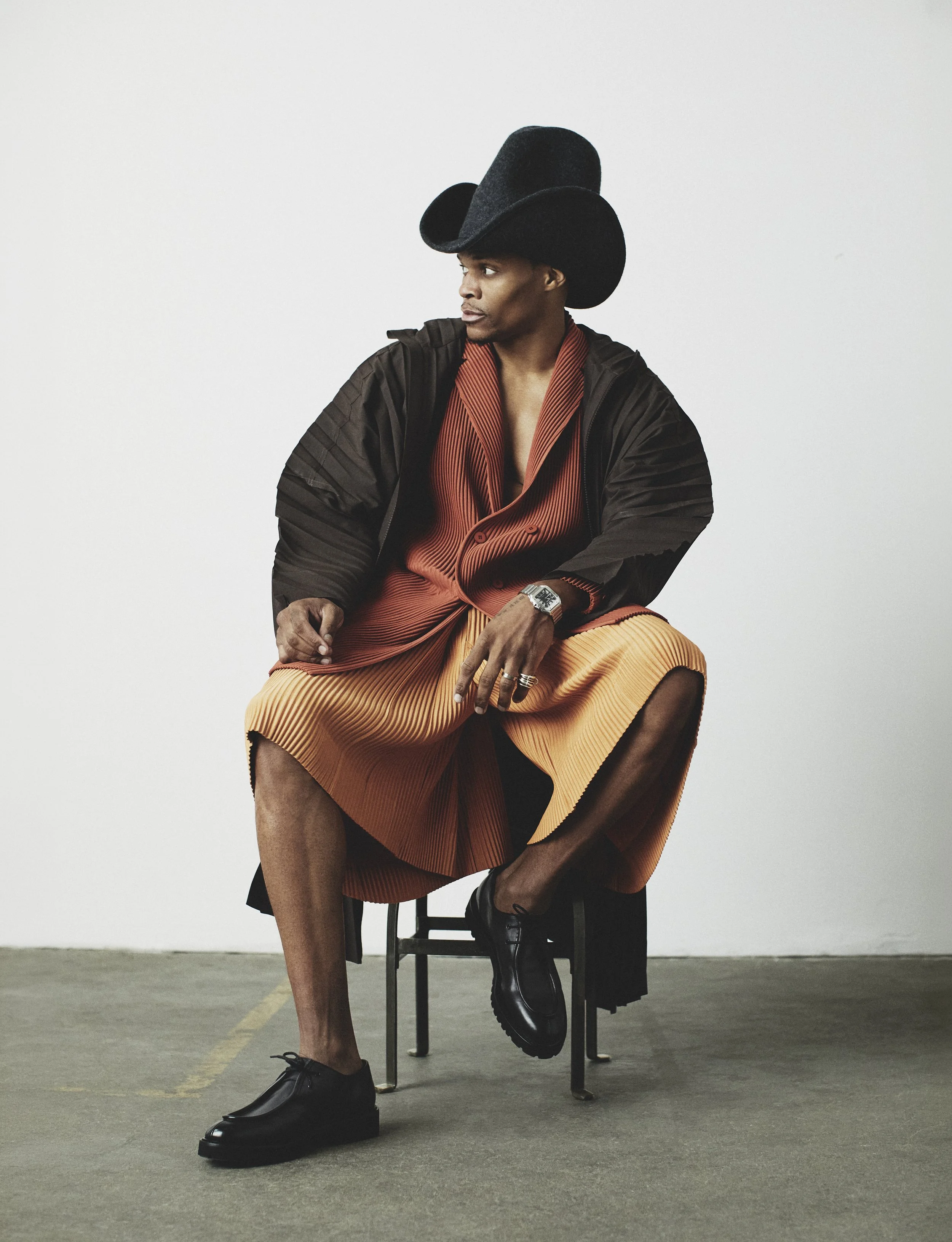Russell Westbrook seated in a sculptural black hat and pleated terracotta outfit against a white studio wall.