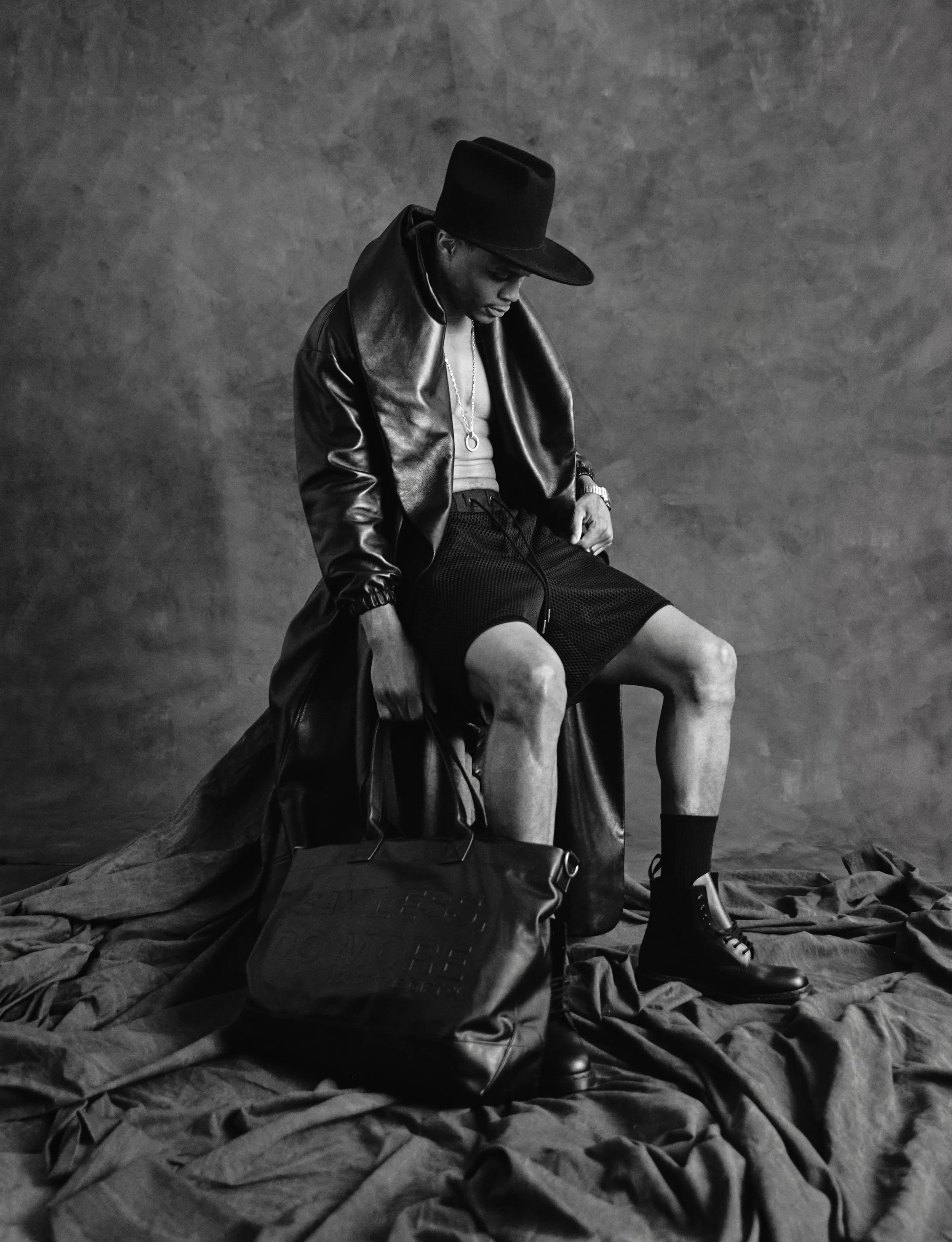 Russell Westbrook seated in an oversized black leather coat and wide-brim hat against a draped canvas backdrop.