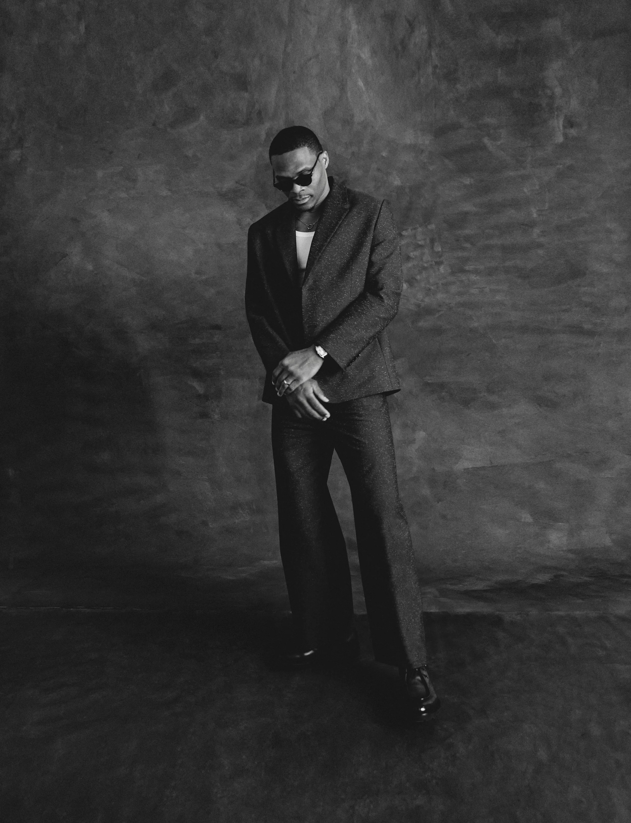 Black and white portrait of Russell Westbrook wearing a dark tailored suit and sunglasses in front of a textured backdrop.