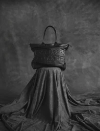 Embossed black leather tote bag reading Say Less Do More resting on a draped fabric pedestal against a textured dark grey backdrop.