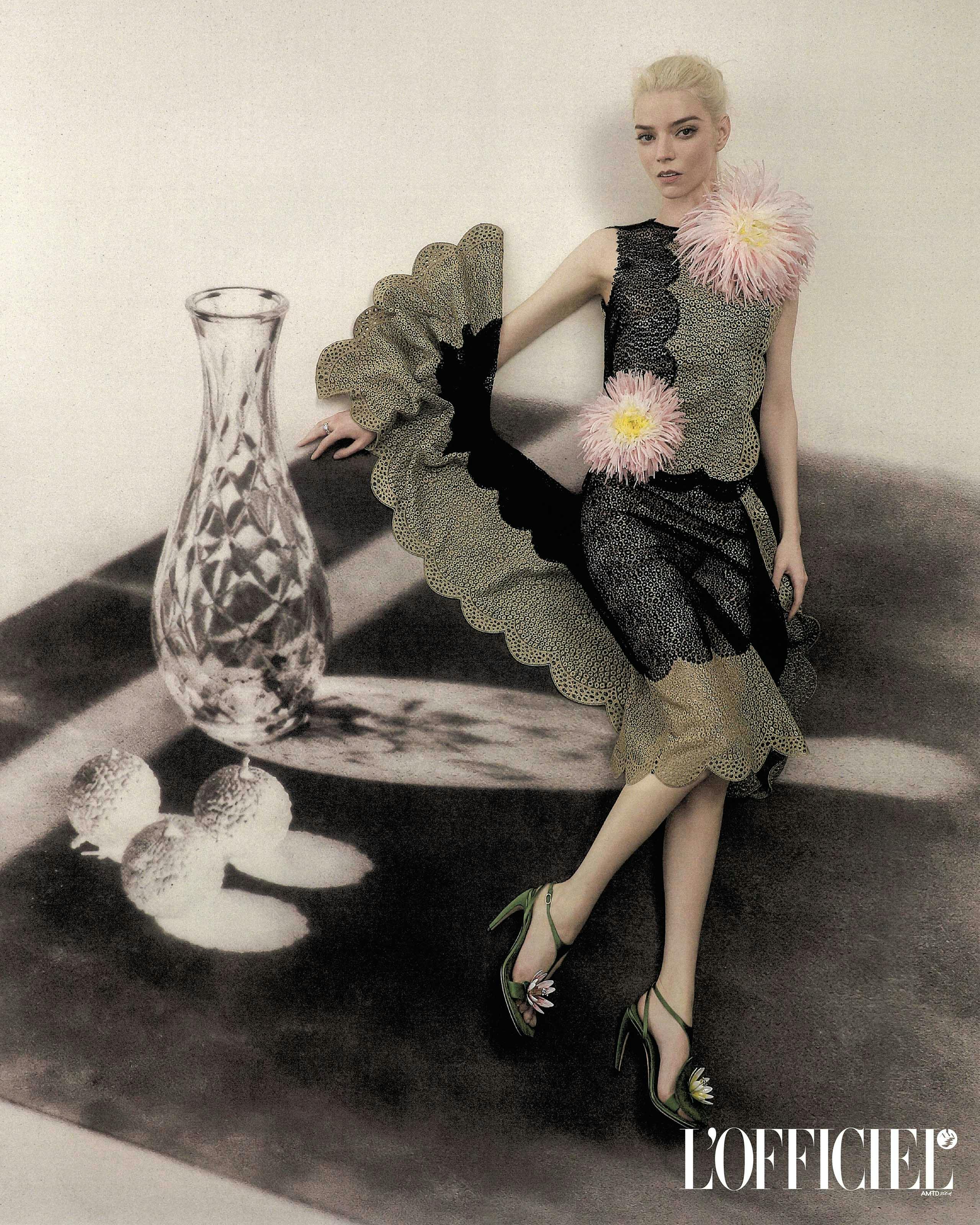 Anya Taylor-Joy in a scalloped black lace dress and green heels posing against a printed still life backdrop.
