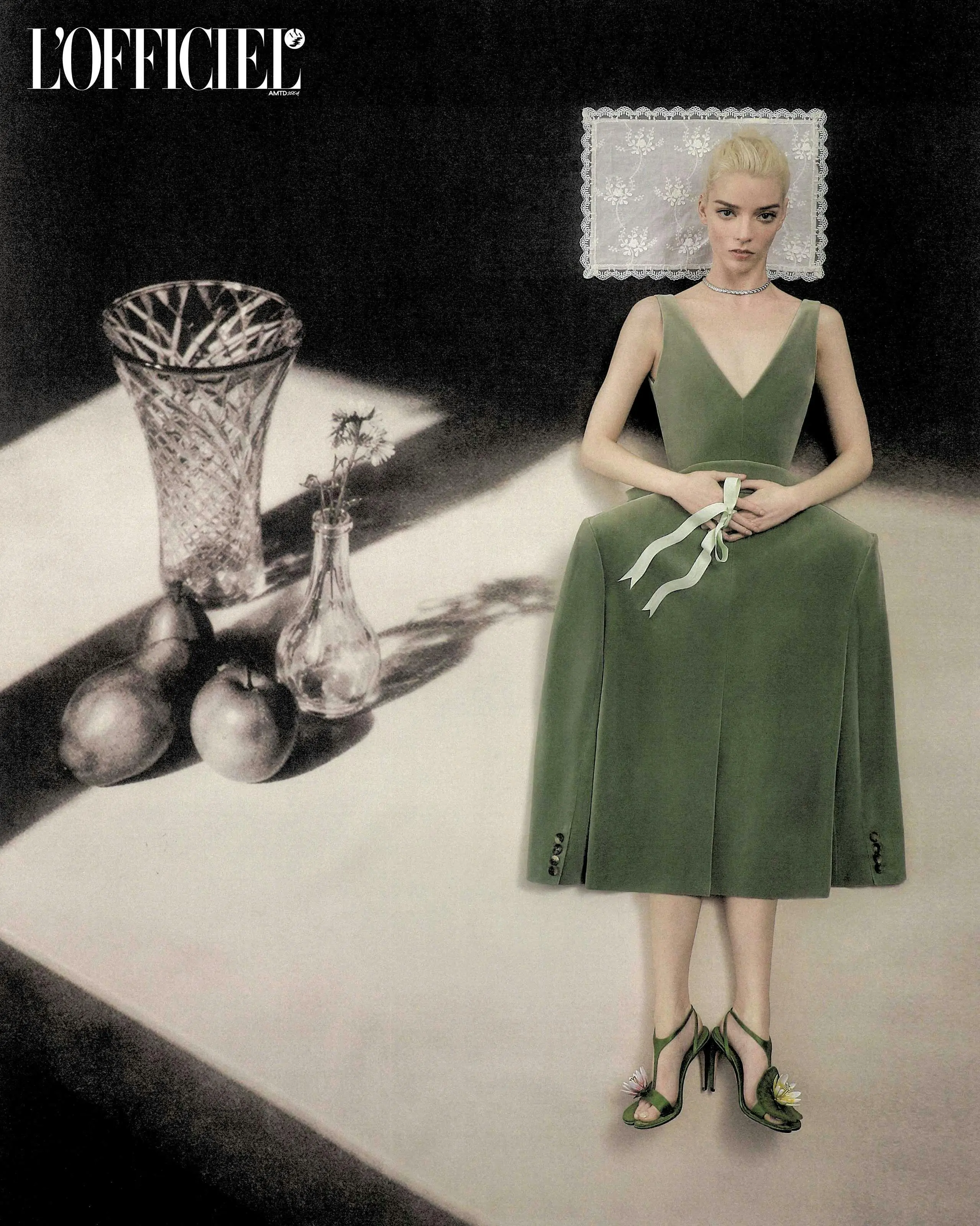 Anya Taylor-Joy wearing a green velvet dress next to a still life arrangement of fruit and vases on a table.