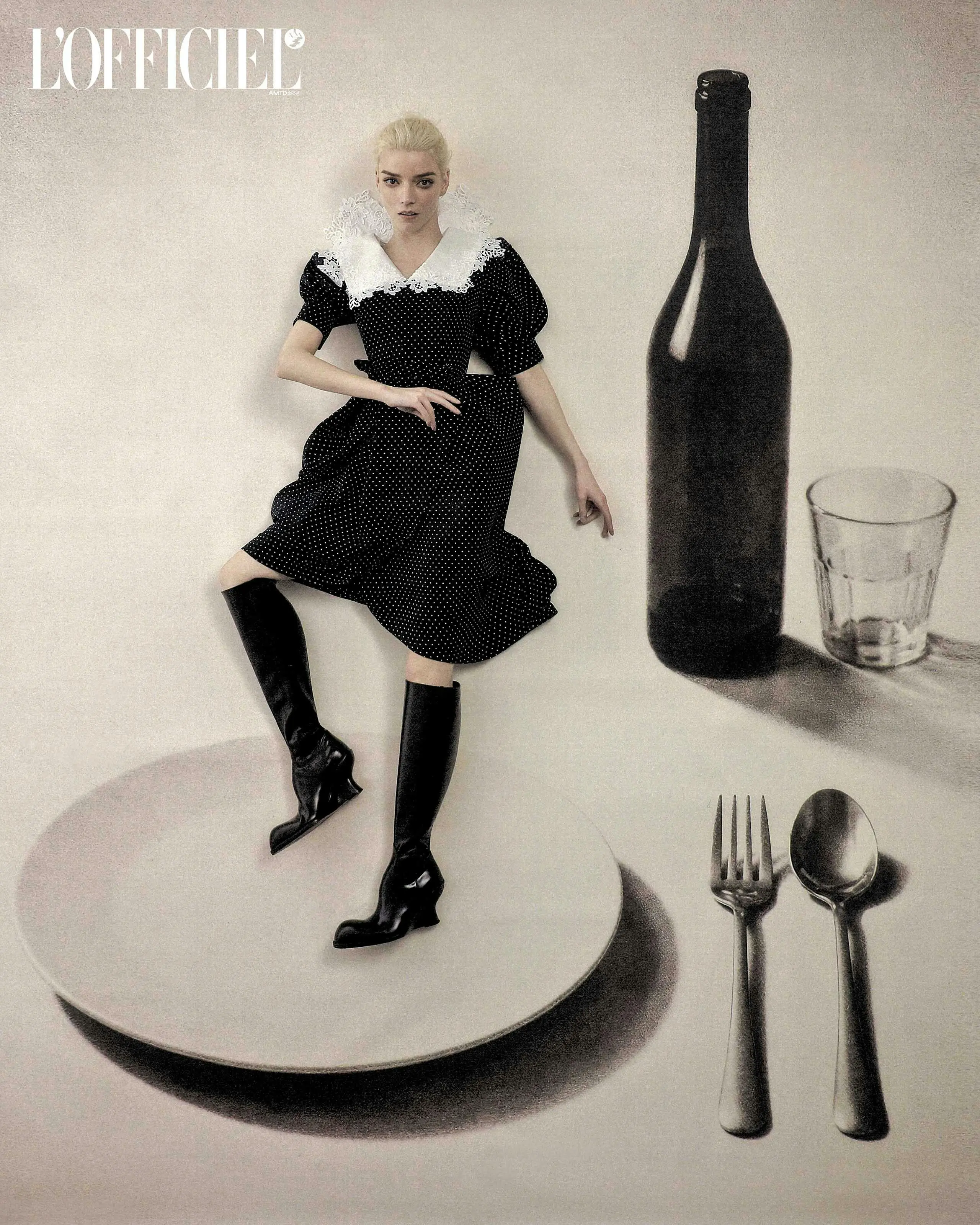Anya Taylor-Joy wearing a black polka dot dress and black boots, posed on a giant dinner plate beside a wine bottle and cutlery.
