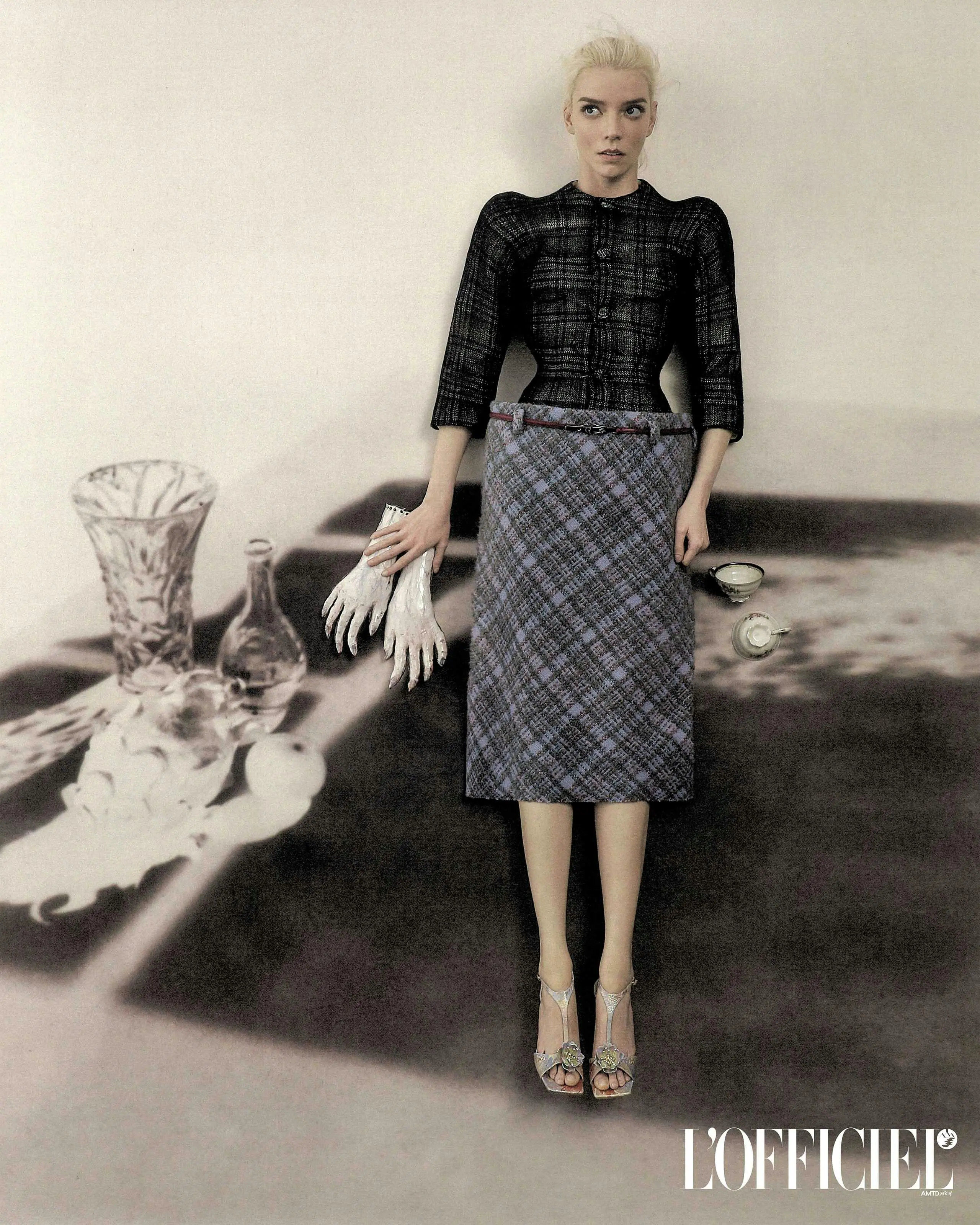 Anya Taylor-Joy wearing a plaid top and skirt standing in a studio with surreal still-life props on a printed floor backdrop.