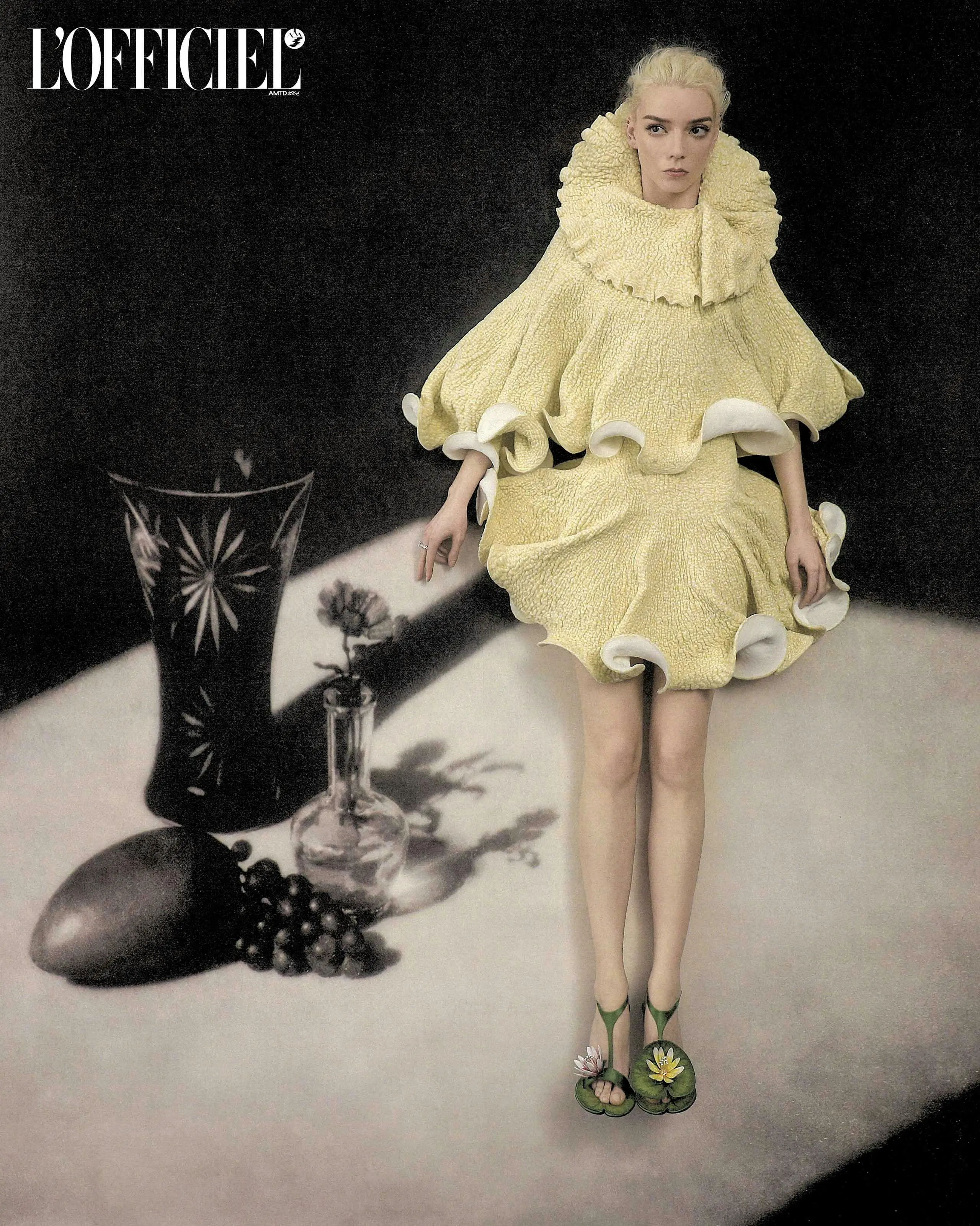 Anya Taylor-Joy posing in a sculptural pale yellow ruffled dress and green floral sandals beside a tabletop still-life arrangement.