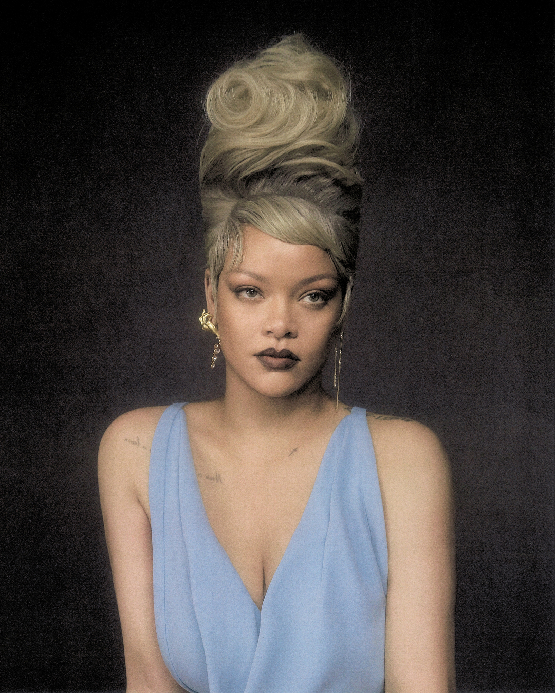 Rihanna with a high sculptural blonde updo and dark lipstick wearing a draped light blue dress against a dark background.