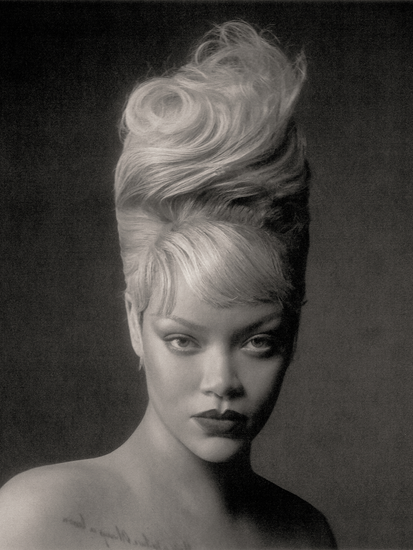 Monochrome close-up portrait of Rihanna featuring a tall sculptural blonde updo and dark lipstick against a dark studio background.