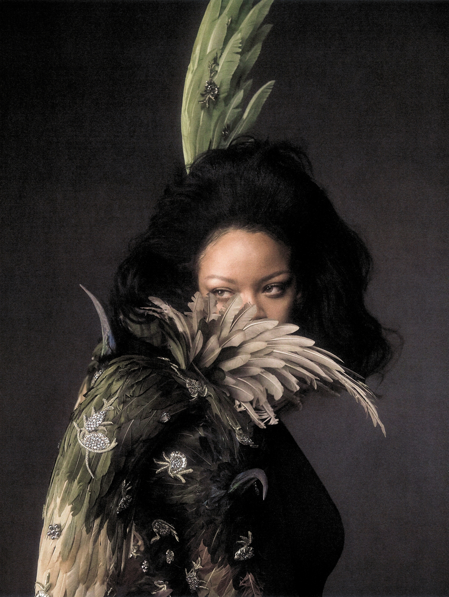 Editorial portrait of Rihanna in a feathered headpiece and embroidered winged cape against a dark textured backdrop.