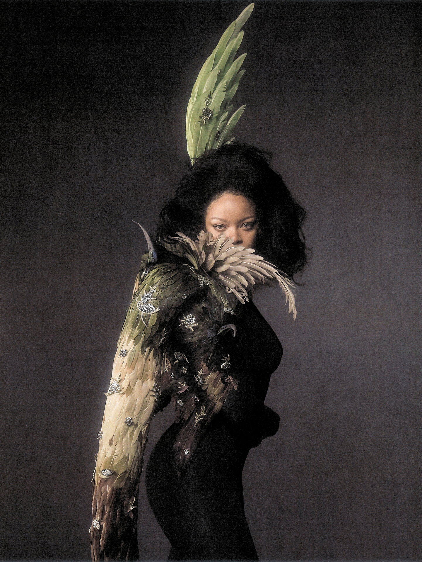 Rihanna wearing feathered sculptural couture with a large feather wing piece, captured in editorial style against a dark textured background.