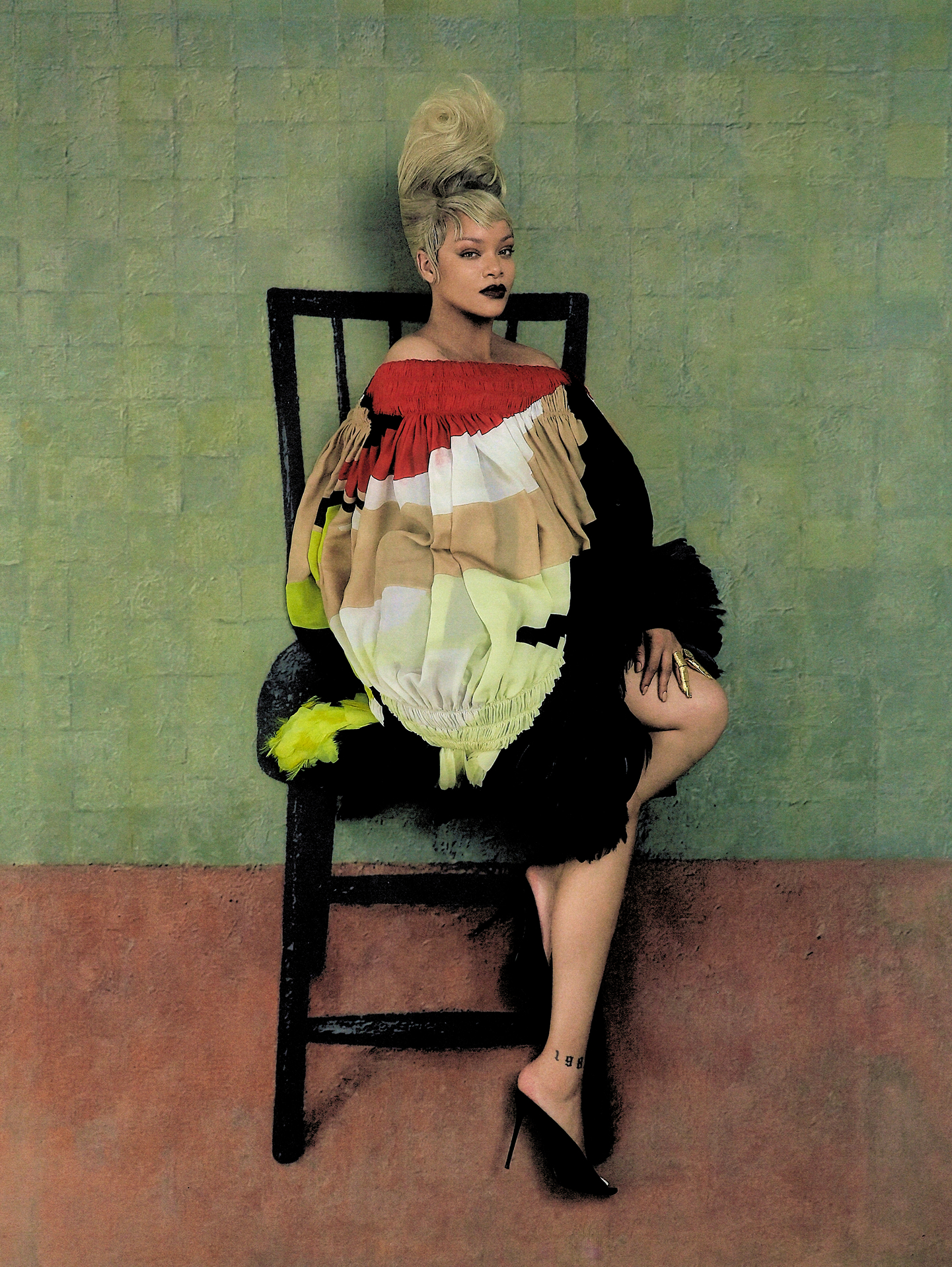 Rihanna seated on a black chair wearing a voluminous multicolored ruffled dress and black pointed-toe heels against a green textured wall.