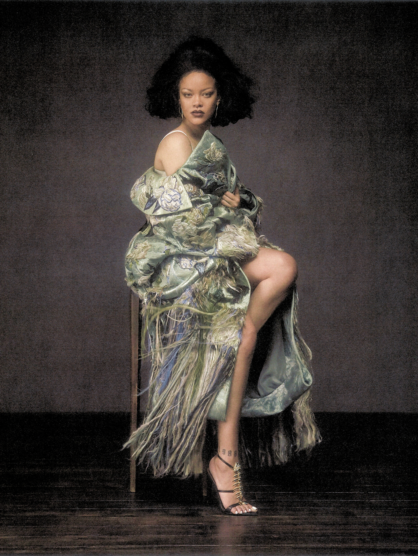 Rihanna seated in an ornate fringed sage green embroidered coat with black strappy heels against a dark textured backdrop.
