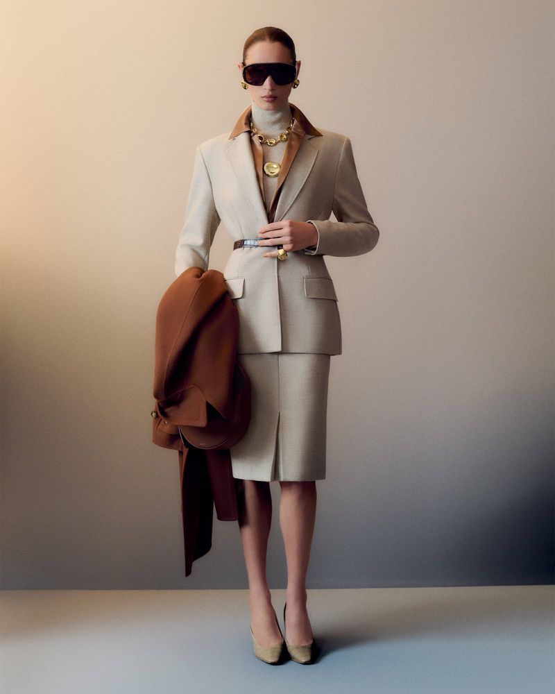 Elegant woman in a beige skirt suit and brown coat, accessorized with gold jewelry and sunglasses, posing in a studio setting.
