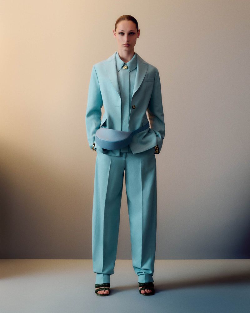 A woman models aqua-colored pantsuit in a studio setting. The outfit includes a jacket, wide-leg pants, and matching belt bag.