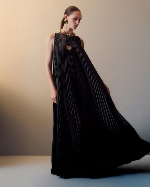 A woman in a flowing, pleated black maxi dress poses in a studio with gold jewelry, including a necklace and earrings, for a fashion shot.