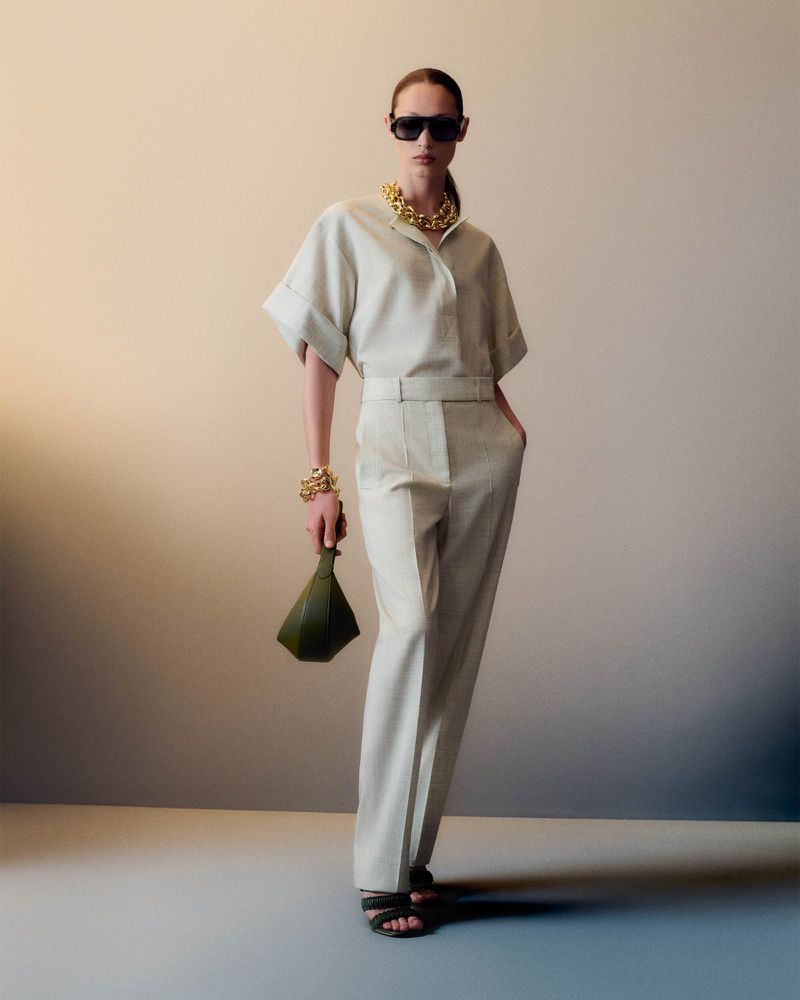 A woman poses in a cream-colored outfit with gold jewelry, holding a green clutch against a neutral backdrop in a fashion shot.