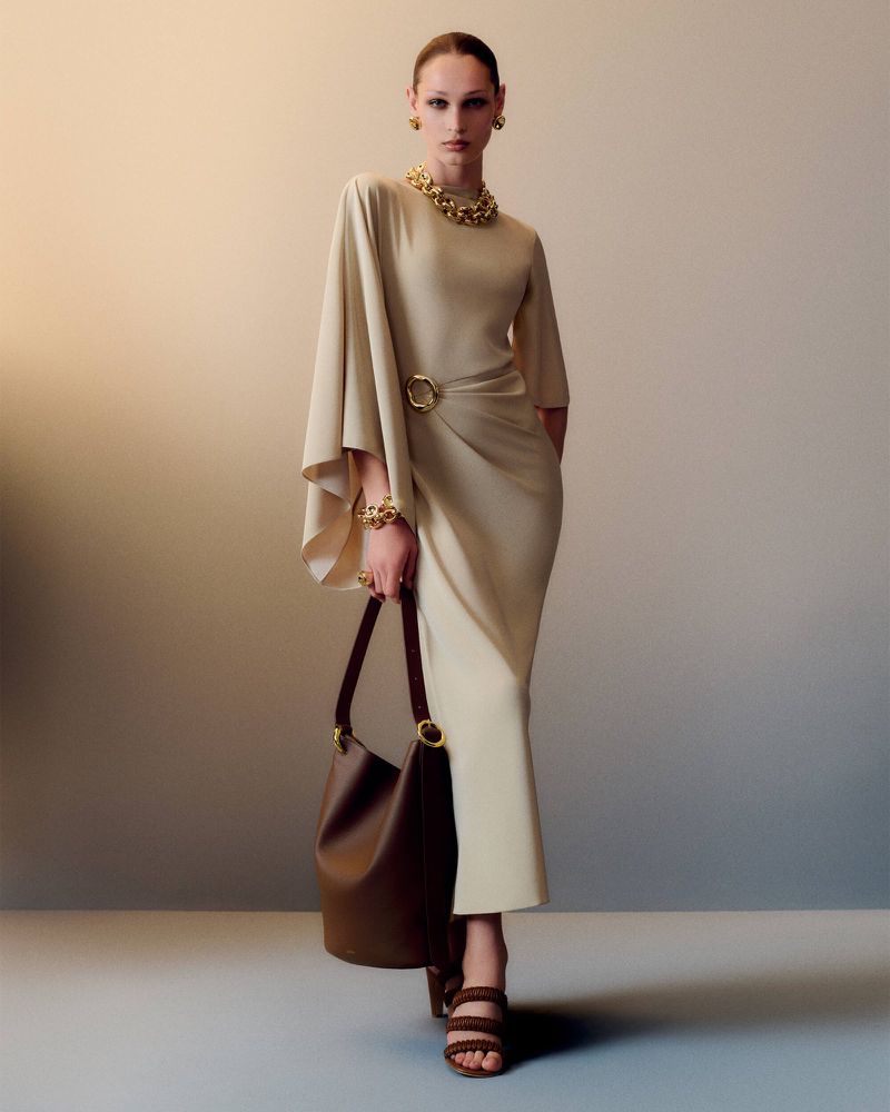 Fashion model poses indoors wearing a light beige dress with gold jewelry and a brown handbag, set against a neutral backdrop.