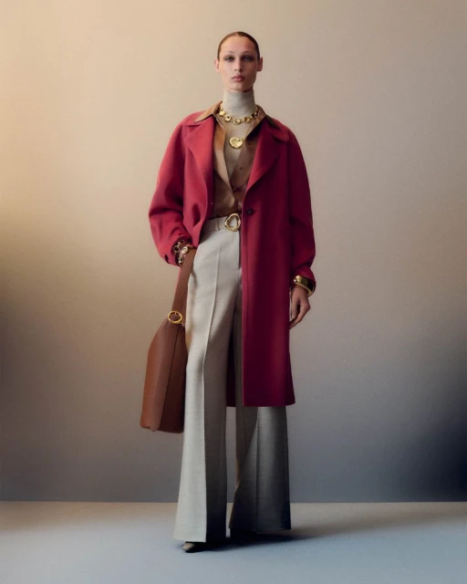 An adult woman poses indoors wearing a pink coat, wide beige pants, a tan turtleneck, gold jewelry, and carrying a brown leather bag.