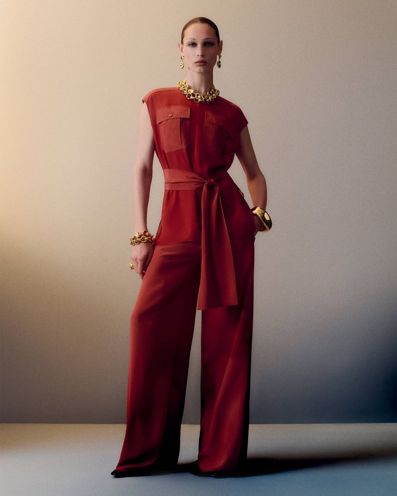 Elegant woman in a red jumpsuit poses with gold jewellery against a neutral backdrop, showcasing a sophisticated fashion style.