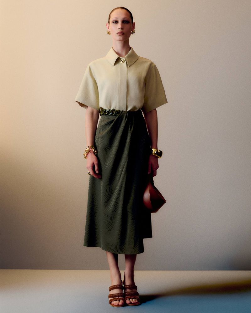 Full-shot of a female model in a collared shirt and wrap skirt, accessorized with jewelry and strappy heels, posing in a studio.