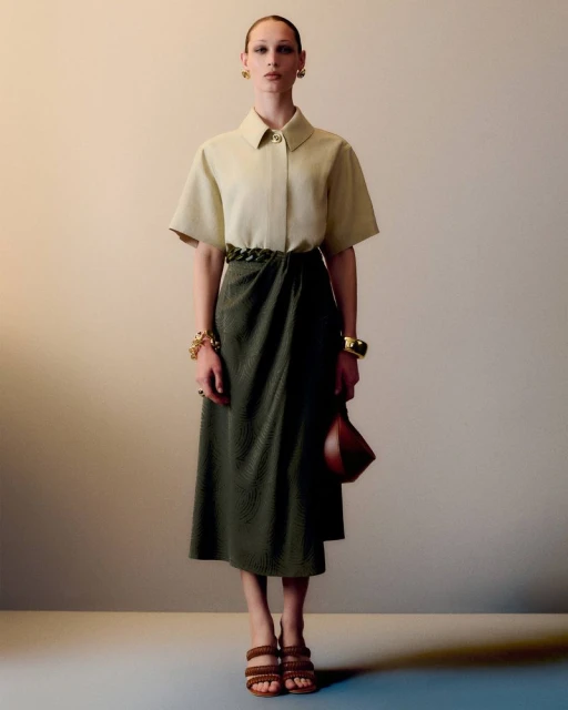 Full-shot of a female model in a collared shirt and wrap skirt, accessorized with jewelry and strappy heels, posing in a studio.
