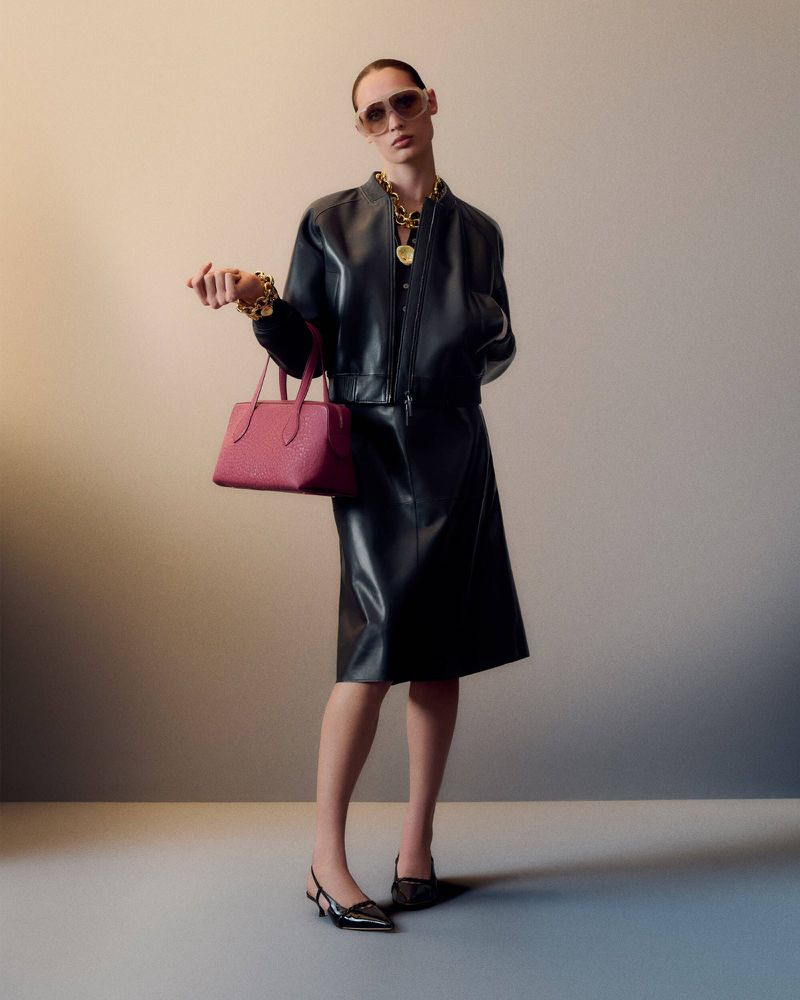 Woman in black leather jacket, skirt, and slingback heels holding a pink handbag and wearing sunglasses and gold jewelry indoors.