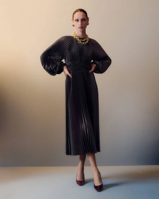 Woman in a pleated midi dress with balloon sleeves, gold necklace and burgundy heels posing in front of a gradient beige background.