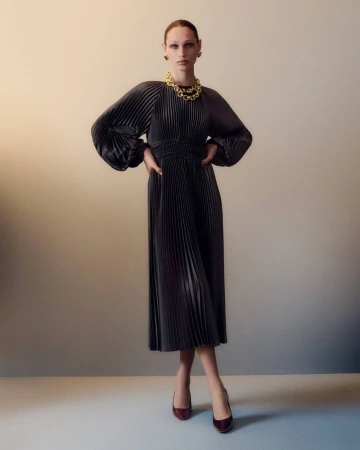 Woman in a pleated midi dress with balloon sleeves, gold necklace and burgundy heels posing in front of a gradient beige background.