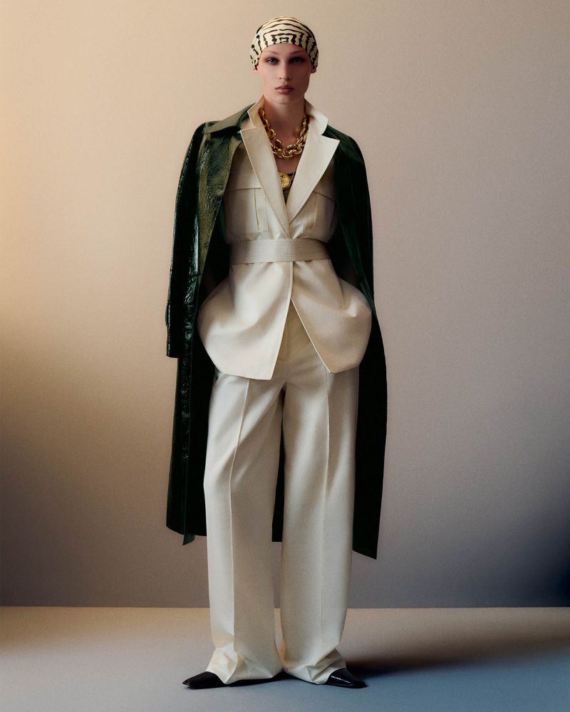 A fair-skinned adult female is featured in a fashion-style full shot, wearing a white belted suit, turban, statement necklace, and patent coat.