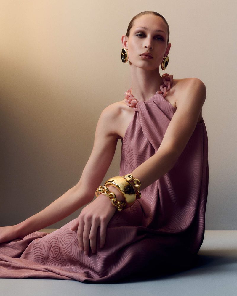 Fashion shot of a fair-skinned female model in a mauve dress with gold bracelets and earrings, set against a neutral-toned background.