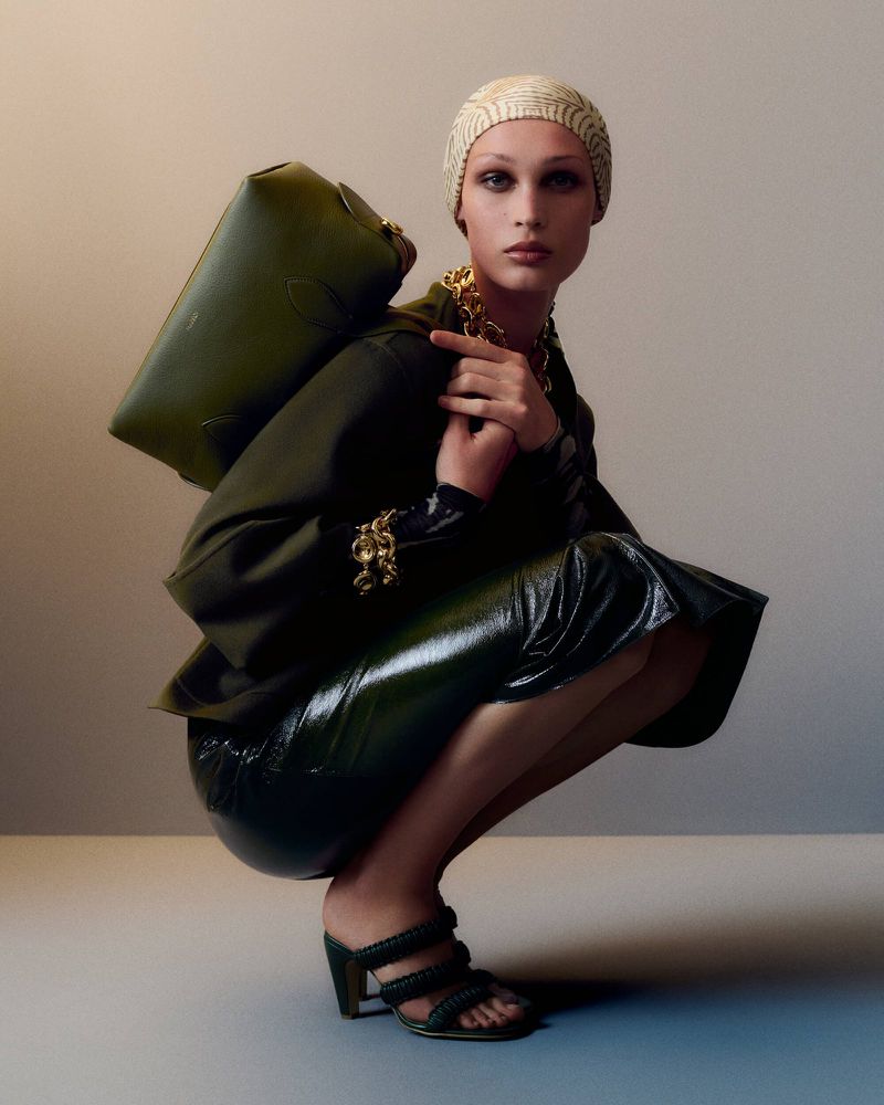 Fashionable adult woman squats in shiny skirt and heels, carrying olive-green handbag and wearing patterned turban and gold jewelry.