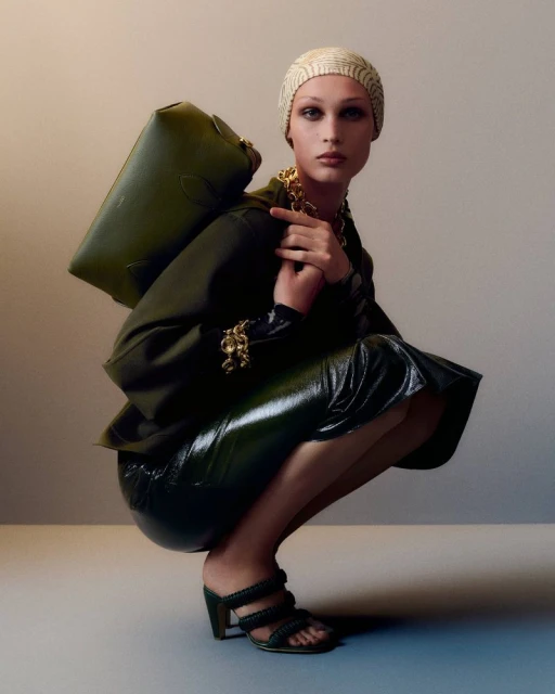 Fashionable adult woman squats in shiny skirt and heels, carrying olive-green handbag and wearing patterned turban and gold jewelry.