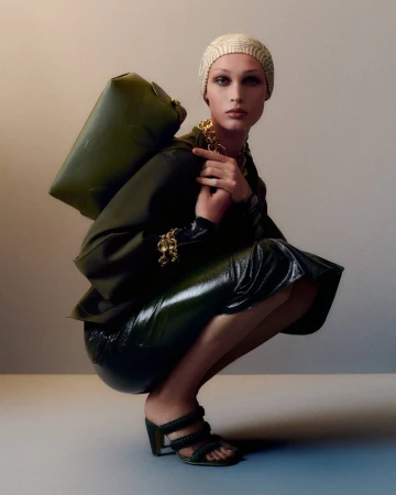 Model Dalton Dubois crouching in an olive green jacket, glossy skirt, and matching shoulder bag against a neutral studio backdrop.